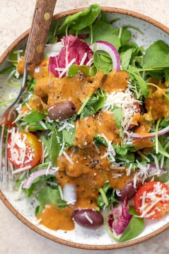 A white bowl with brown rim filled with salad greens with tomatoes and olives tossed with sun dried tomato vinaigrette. A fork with a brown wood handle sits in the bowl.