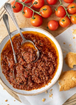 A white bowl filled with sun-dried tomato pesto. Two spoons sit in the bowl and pieces of baguette sit next to the bowl.