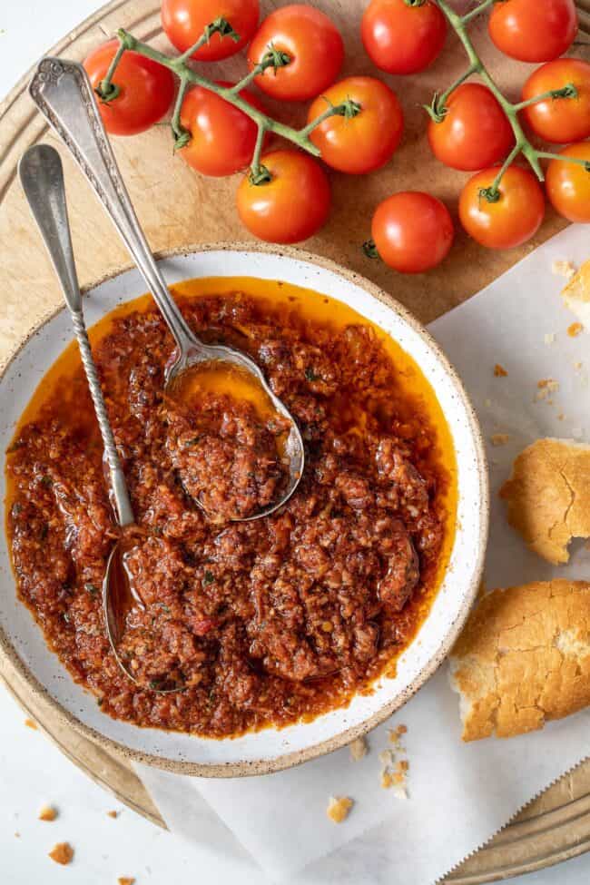 A white bowl filled with red pesto. Two spoons sit in the red pesto sauce with pieces of bread sit next to the bow.