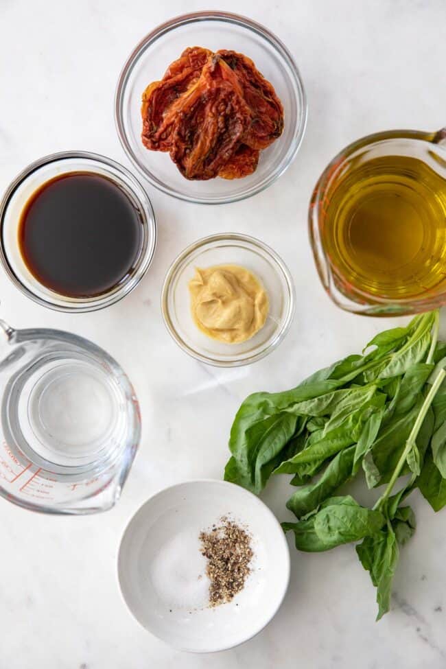 Glass bowls filled with olive oil, balsamic vinegar, sun dried tomatoes and water. Fresh basil leaves sits next to the bowls.