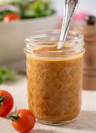 A mason jar with sun dried tomato vinaigrette with a spoon in it. A white bowl filled with salad sits next to the mason jar.