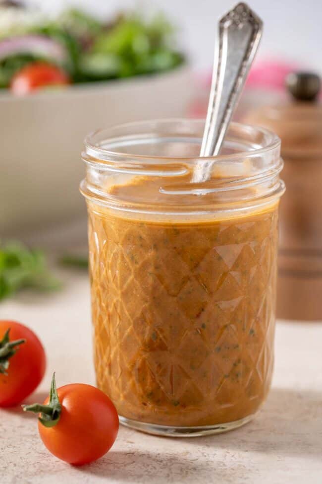 Mason jar filled with sun dried tomato vinaigrette. A white bowl filled with salad sits next to the mason jar.