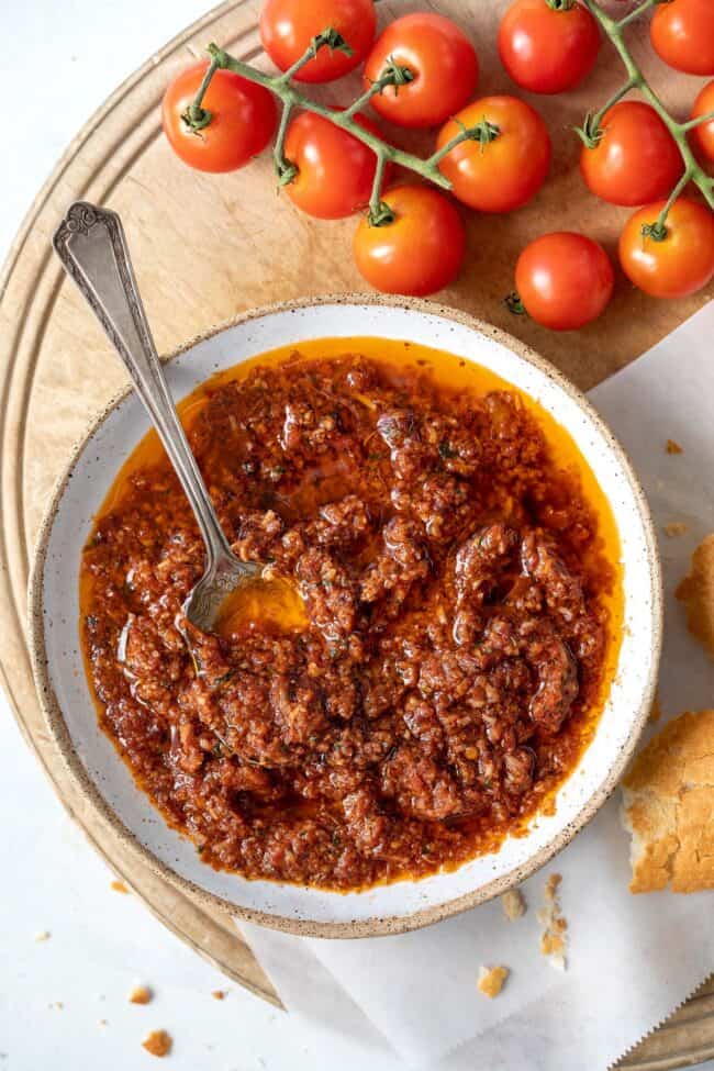 A whit bowl filled with sun dried tomato pesto (red pesto sauce). A spoon sits in the bowl and crusty bread sits next to the bowl.