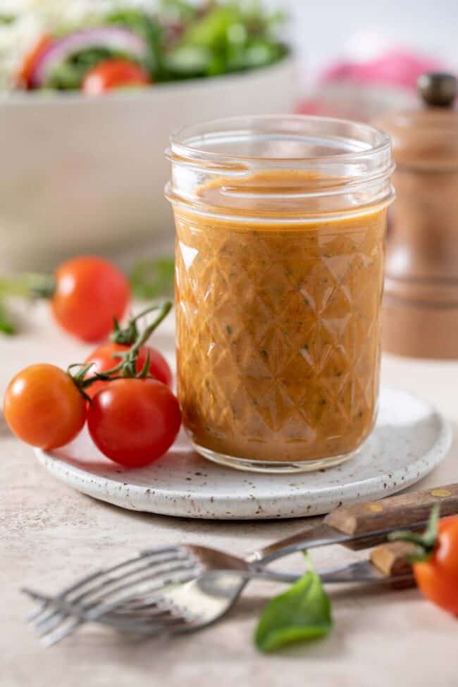 A mason jar filled with salad dressing. A white bowl filled with salad and two forks with brown wooden handles sits next to the mason jar.