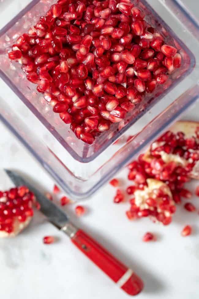 A blender filled with pomegranate seeds. A red knife and broken pieces of pomegranate sit next to the blender.