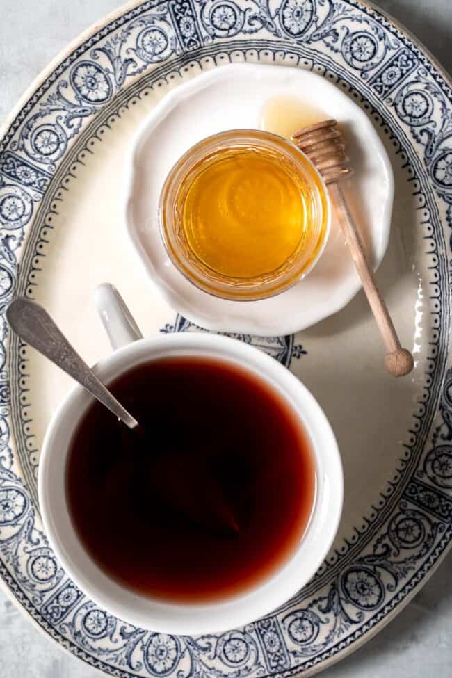 A white cup filled with elderberry tea and a glass jar filled with honey sit on a blue and white oval platter.