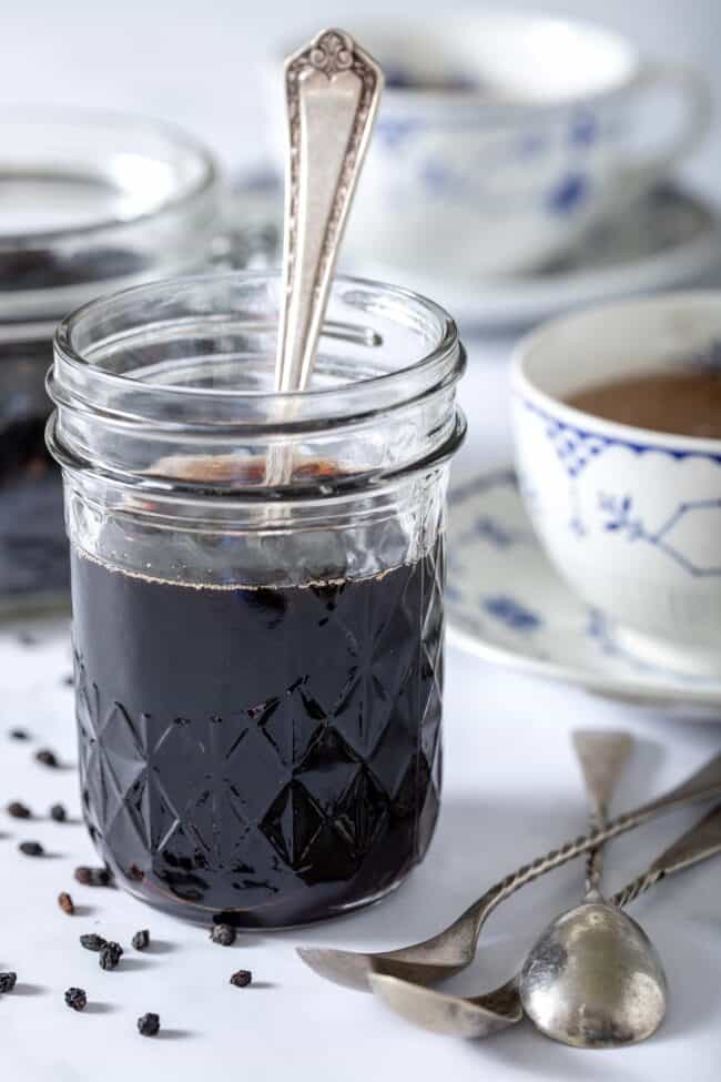 A clear glass mason jar filled with elderberry syrup. A white and blue teacup sits next to the mason jar.