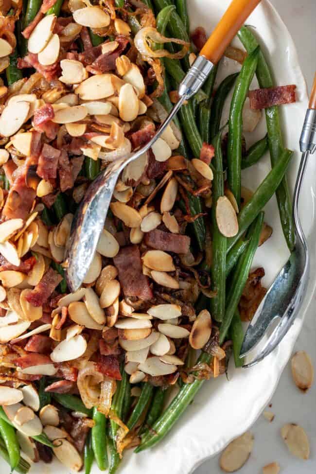 White platter filled with haricot vert (French green beans) covered with chopped turkey bacon and toasted almonds. A silver and yellow server sits on the platter.