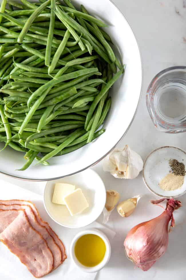 Large white bowl filled with green beans. Next to the bowl sit slices of turkey bacon, a cup of water, garlic, shallot, and a small white dish with butter slices.