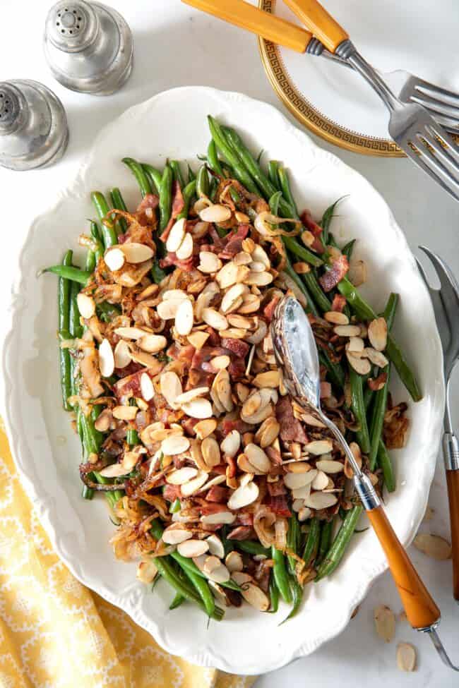 White platter filled with haricot vert (French green beans) covered with chopped turkey bacon and toasted almonds. A silver and yellow server sits on the platter.
