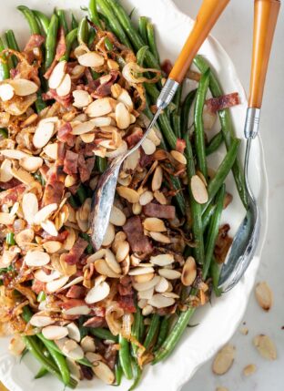 White platter filled with haricot vert (French green beans) covered with chopped turkey bacon and toasted almonds. A silver and yellow server sits on the platter.