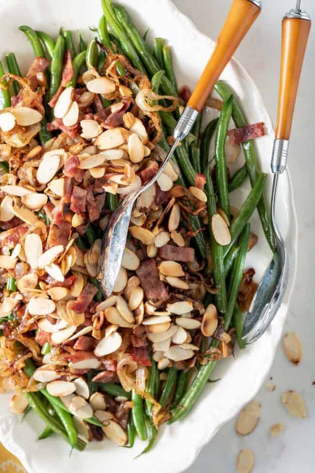 White platter filled with haricot vert (French green beans) covered with chopped turkey bacon and toasted almonds. A silver and yellow server sits on the platter.