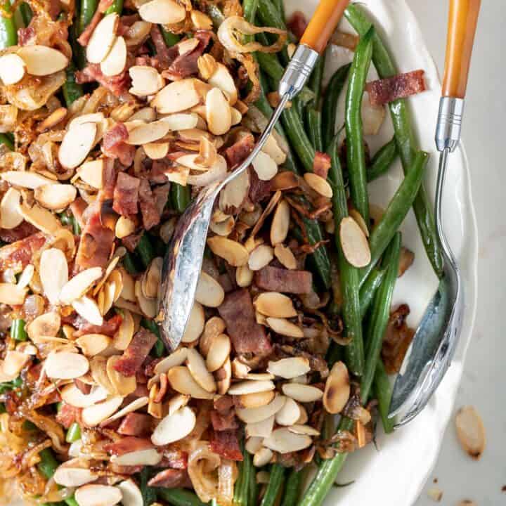 White platter filled with haricot vert (French green beans) covered with chopped turkey bacon and toasted almonds. A silver and yellow server sits on the platter.