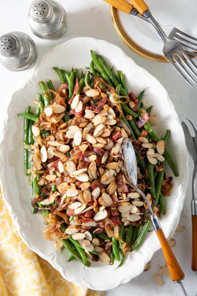 White platter with green beans and toasted almonds. Serving tongs rest on the platter with a yellow napkin sitting next to the platter.