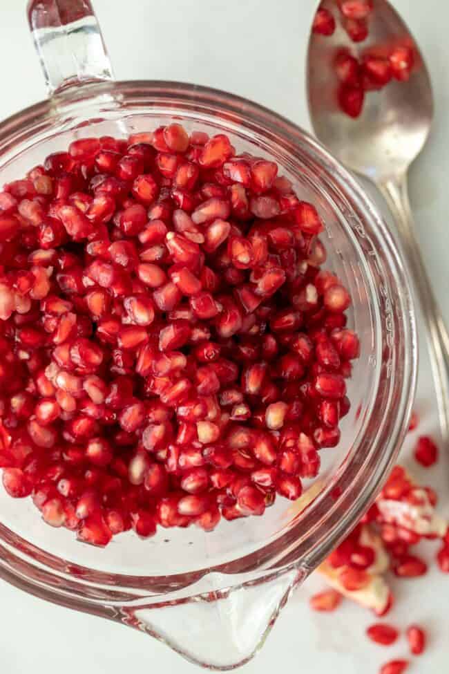 A clear glass measuring cup filled with red fruit seeds. Scattered seeds sit next to the cup.