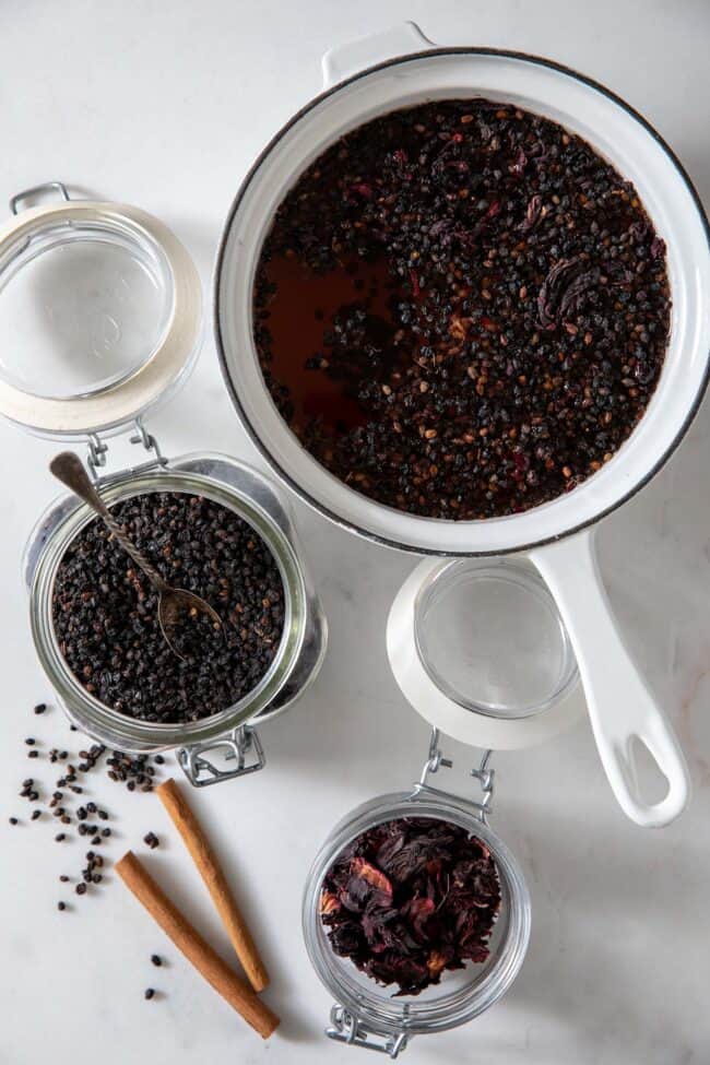 White pot filled with elderberry tea. Two mason jars filled with dried elderberries and hibiscus sit next to the pot.