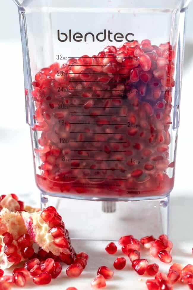 A clear blender pitcher filled with pomegranate seeds. Seeds are scattered on the counter next to the blender.