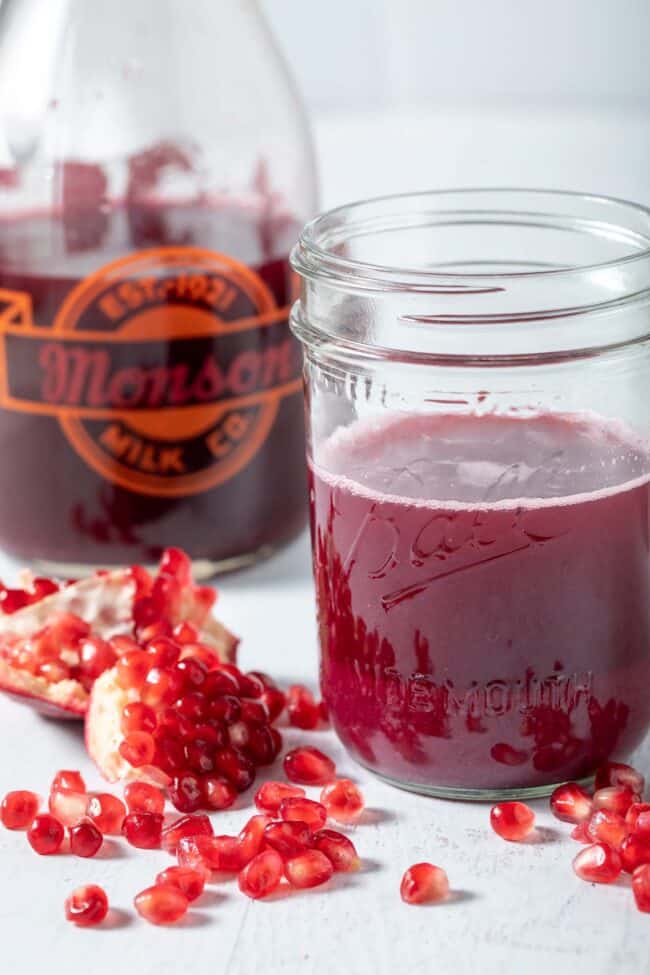 A glass pitcher filled with pomegranate juice sits next to a mason jar filled with pomegranate juice. Pomegranate seeds are scattered next to the mason jar.