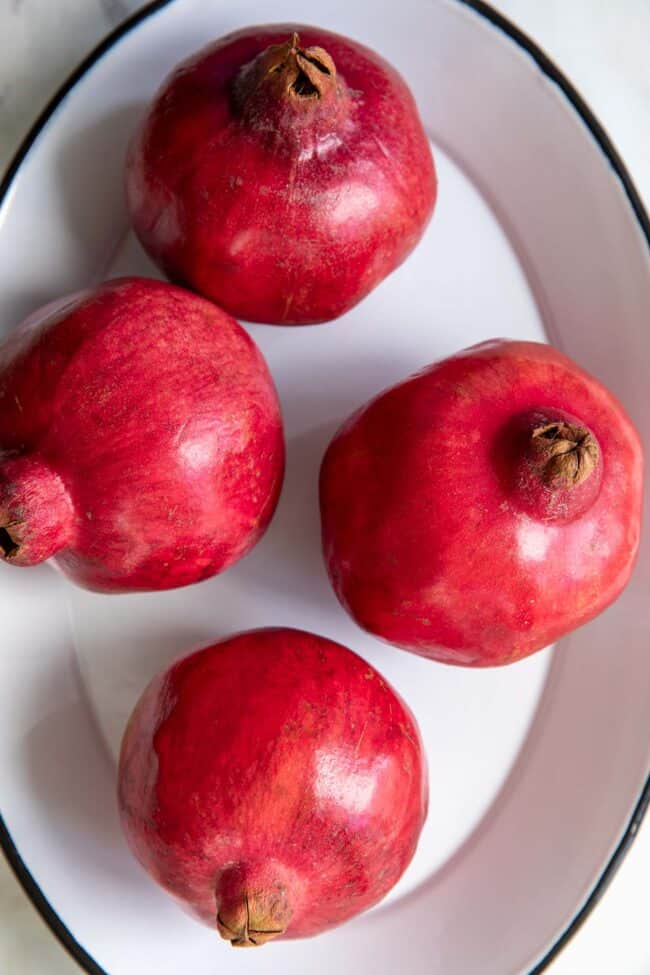 Four whole pomegranates sitting on a white oval metal platter with black rim.