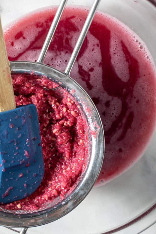 A clear glass bowl is filled with pomegranate juice. A strainer with a blue silicon spatula and pomegranate seeds sits over the glass bowl.