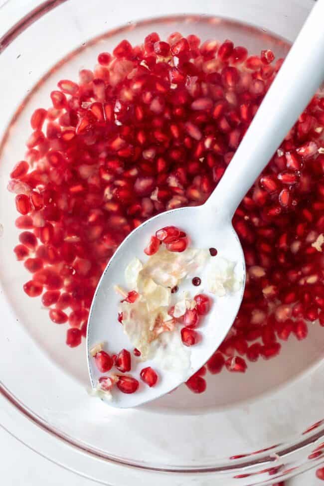 A glass bowl filled with water and pomegranate seeds sit at the bottom. A white straining spoon rests in the water with a few pomegranate seeds.