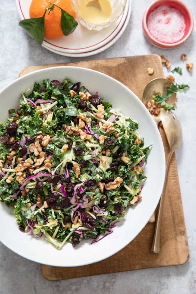 White bowl filled with chopped kale salad with cranberries and walnuts. Gold serving spoons sit next to the bowl.