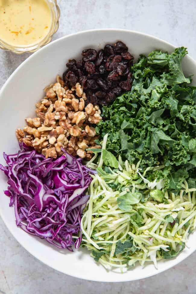 A white bowl filled with chopped kale, broccoli slaw, purple cabbage, walnuts and dried cranberries. A clear glass cup with orange dressing sits next to the bowl.