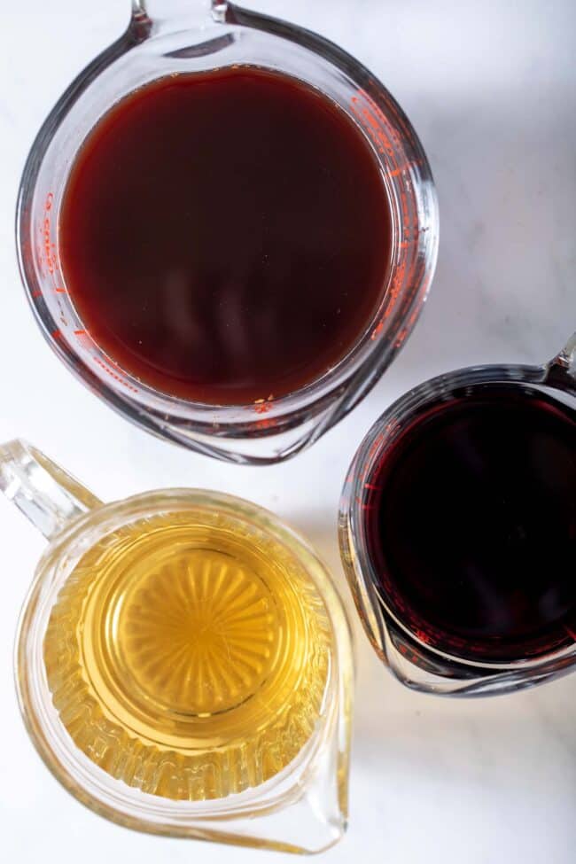 Three clear glass measuring cups filled with pomegranate juice, elderberry tea and white grape juice.