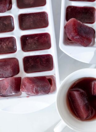 A white ice cube tray is filled with elderberry pomegranate ice cubes. A white tea cup with two melting pomegranate ice cubes sits next to the tray.