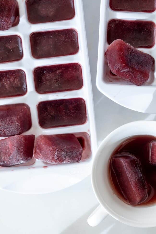 white ice cube tray filled with purple ice cubes. A white tea cup next to the ice cube tray has 2 melting purple ice cubes in it