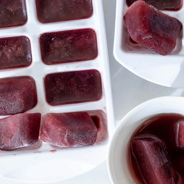 A white ice cube tray is filled with elderberry pomegranate ice cubes. A white tea cup with two melting pomegranate ice cubes sits next to the tray.