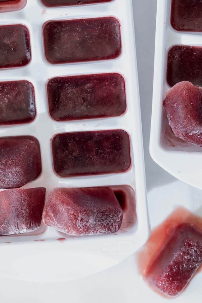 Two white ice cube trays filled with pomegranate and elderberry ice cubes (frozen elderberry tea cubes). 