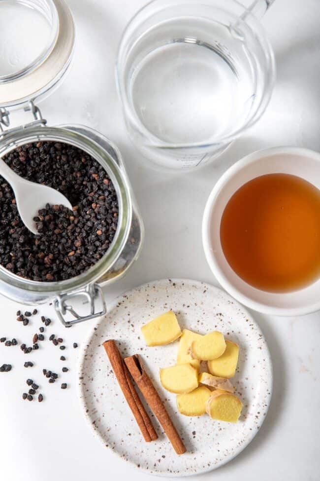 A measuring cup filled with water, a white bowl filled with honey, a jar of dried elderberries and a dish with cinnamon stick and slices of giner.