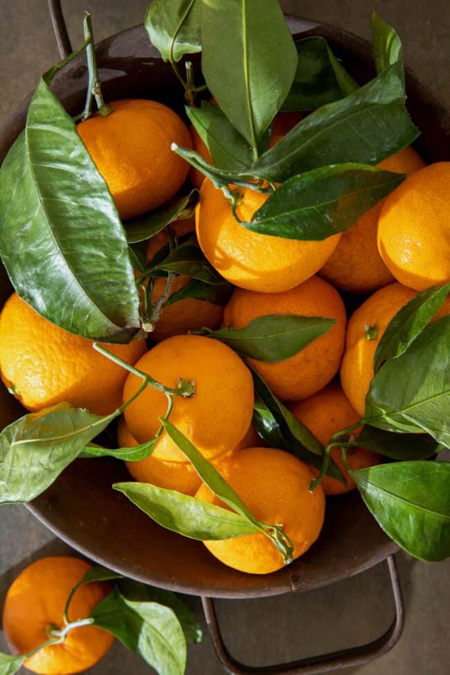 A brown colander filled with mandarin oranges with their green leaves attached.