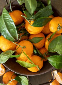 A brown colander filled with mandarin oranges with their green leaves attached. A peeled mandarin orange sits next to the colander.