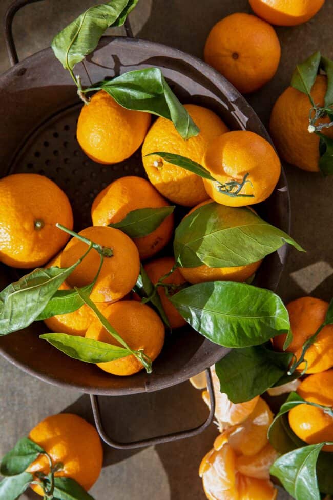 A brown colander filled with citrus fruit with their green leaves attached.