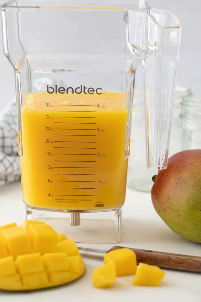 Clear plastic blender pitcher filled with blended juice. A cut mango and knife sits next to the pitcher.