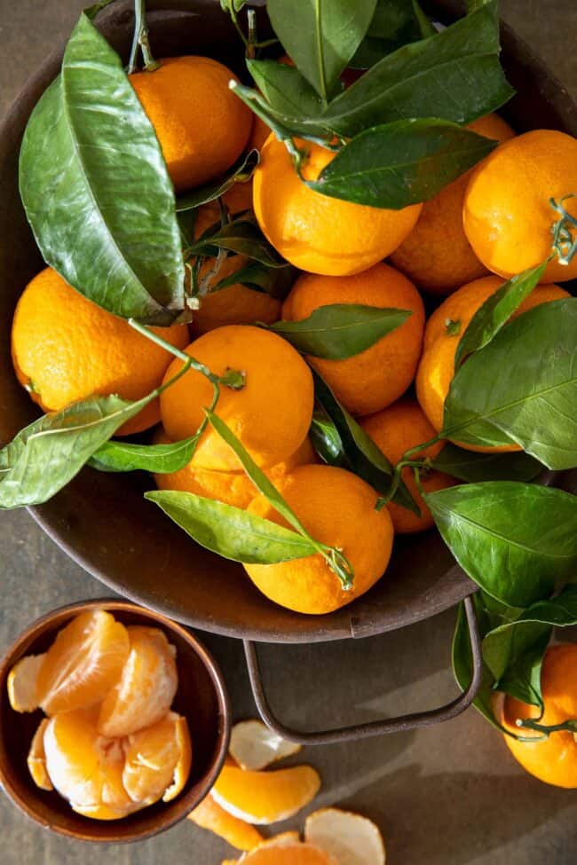 A brown colander filled with clementine oranges with their green leaves attached. A peeled clementine orange sits next to the colander.