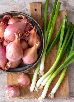 Brown cutting board with bowl of shallots. Green onions sit loosely on the cutting board next to the bowl.