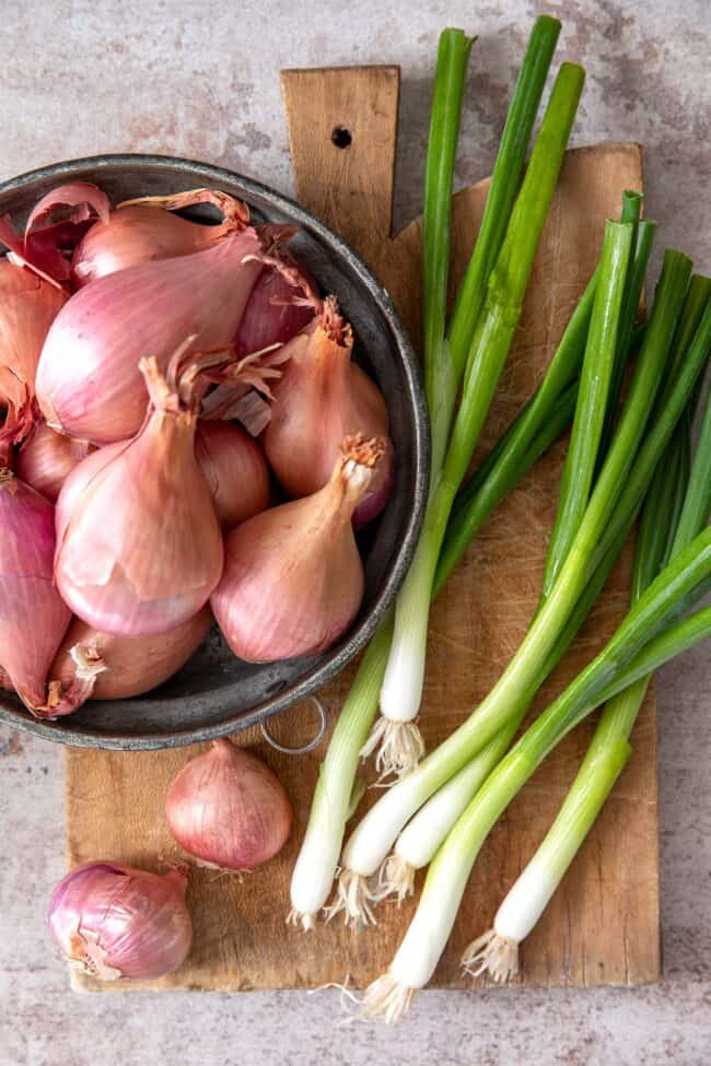 Brown cutting board with bowl of shallots. Green onions sit loosely on the cutting board next to the bowl.