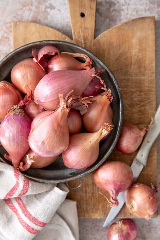 A metal bowl filled with shallots sits on a wooden cutting board. A few loose shallots and a knife sits next to the bowl.