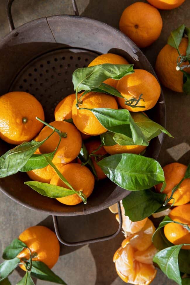 A brown colander filled with mandarin oranges with their green leaves attached. A peeled mandarin orange sits next to the colander.