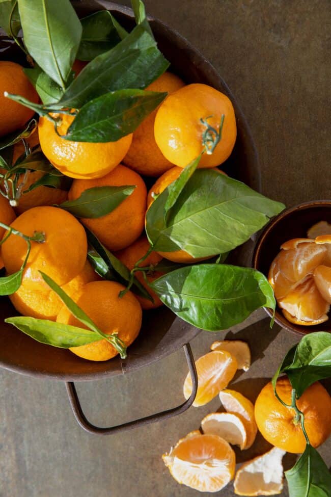 A brown colander filled with satsuma oranges with their green leaves attached. A peeled satsuma orange sits next to the colander.