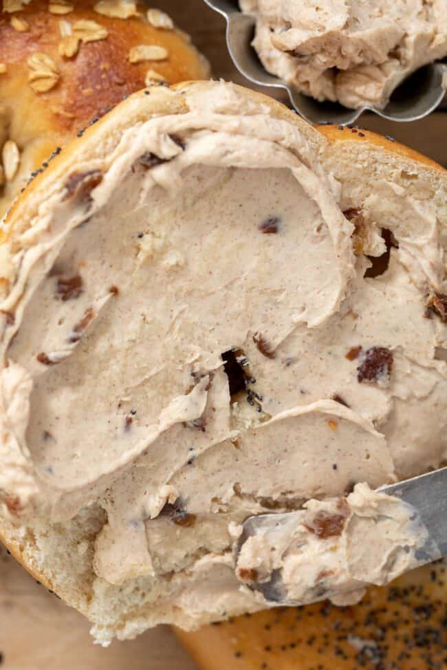 A bagel with cinnamon raisin cream cheese slathered on it with a knife.