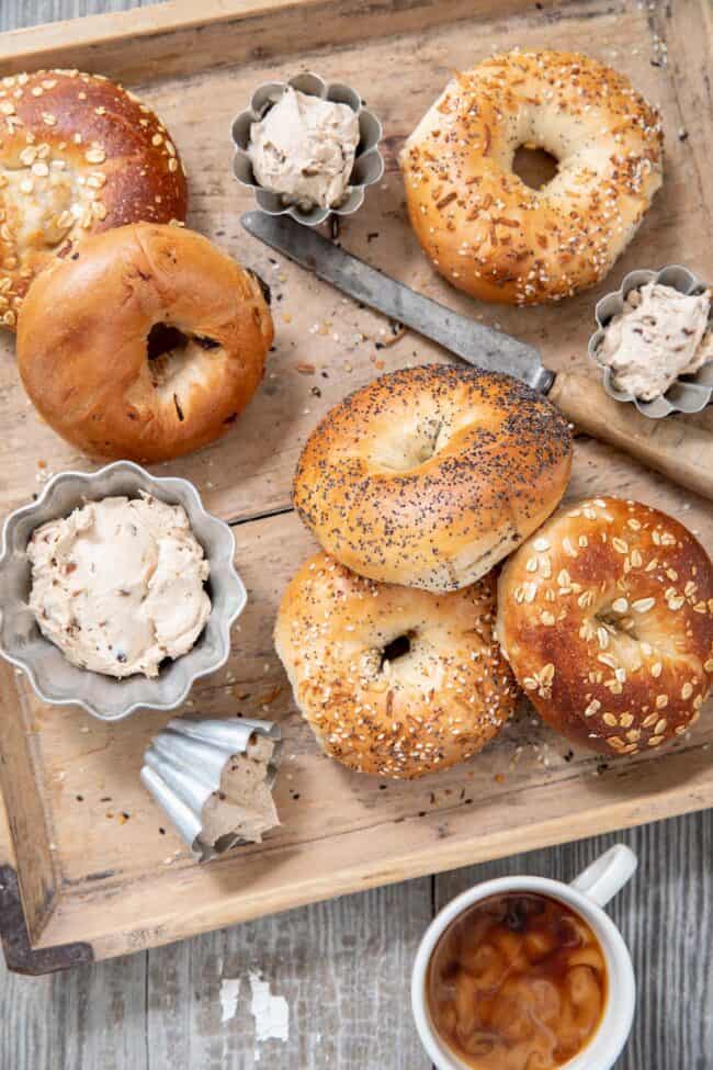 Bagels scattered on a wooden tray with silver ramekins filled with a creamy spread for the bagels.