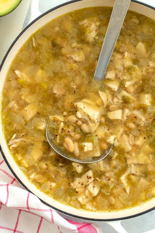 A white pot filled with white bean chicken chili. A silver ladle sits in the pot and a red and white dish towel sits next to the pot.