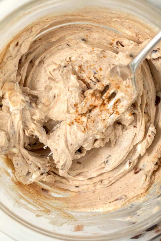 A clear glass bowl with blended cream cheese with cinnamon and raisins. A fork is in the middle of the bowl with the mixture on it.