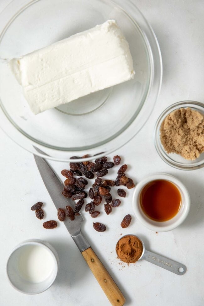 Glass mixing bowl with a block of cream cheese. Small bowls filled with brown sugar, pure vanilla extract, milk and cinnamon sit next to the bowl. Raising scattered over a knife also sits next to the bowl.