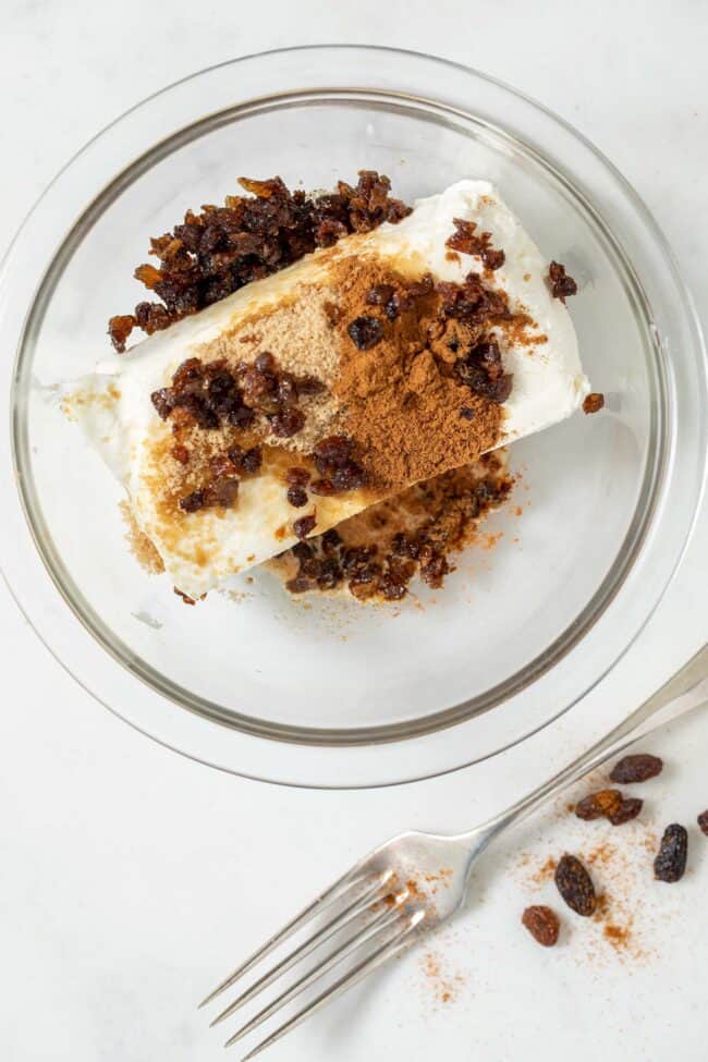 A clear glass mixing bowl is filled with a block of cream cheese, ground cinnamon, brown sugar and chopped raisins. A silver fork sits next to the bowl.