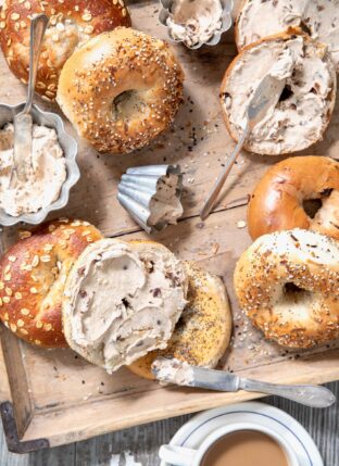 Bagels scattered on a wooden tray with silver ramekins filled with cinnamon raisin cream cheese spread.